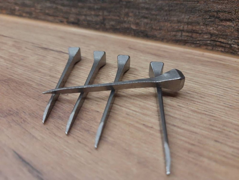 May include: Five silver horseshoe nails are arranged on a wooden surface. The nails are made of metal and have a rectangular head and a pointed tip. These nails are used to attach horseshoes to a horse's hoof.