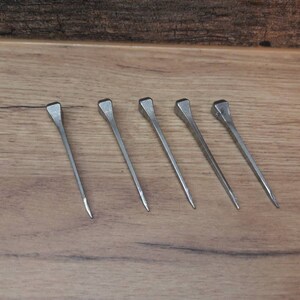 May include: Five silver nails with square heads are arranged on a wooden surface. The nails are evenly spaced and angled slightly to the right. The wood grain is visible in the background.