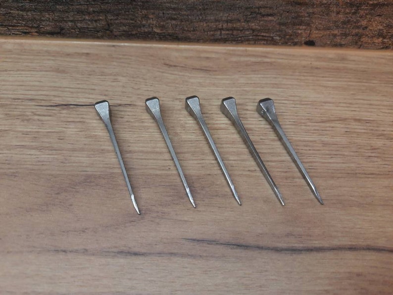 May include: Five silver nails with square heads are arranged on a wooden surface. The nails are evenly spaced and angled slightly to the right. The wood grain is visible in the background.