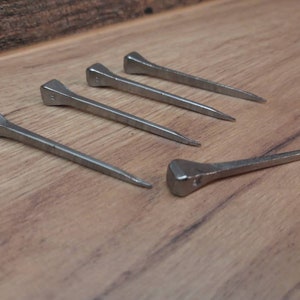 May include: Five silver horseshoe nails with square heads and tapered points are arranged on a wooden surface. The nails are evenly spaced, with one nail positioned separately from the others.