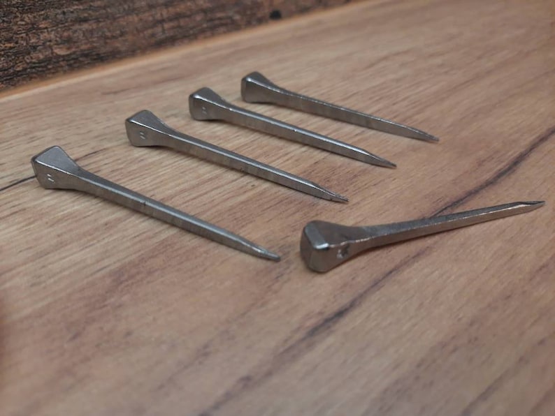 May include: Five silver horseshoe nails with square heads and tapered points are arranged on a wooden surface. The nails are evenly spaced, with one nail positioned separately from the others.