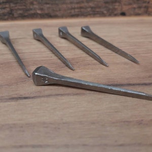 May include: Five silver horseshoe nails on a wooden surface. The nails have a rectangular head and a tapered, pointed end. The wood grain is visible in the background.
