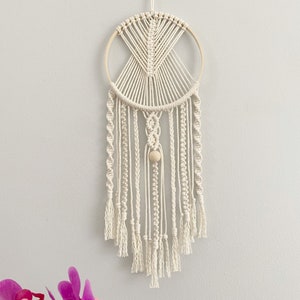 May include: Cream-colored macrame wall hanging featuring a circular wooden hoop and intricate woven patterns. The design includes a central geometric shape, vertical strands, and a wooden bead, creating a bohemian aesthetic. The bottom has a fringe.