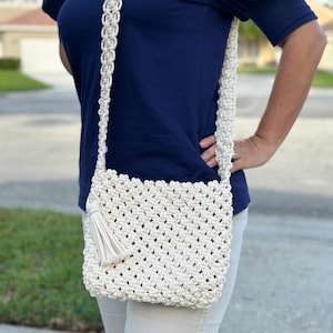 Puede incluir: Bolso de hombro de macramé color crema con un patrón tejido y una borla. El bolso tiene una correa larga y se lleva cruzado. El bolso está hecho de una fibra natural suave.