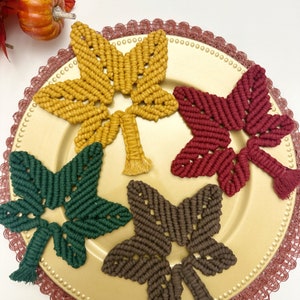 Puede incluir: Cuatro posavasos de macramé hechos a mano en diferentes colores: amarillo mostaza, rojo burdeos, verde bosque y marrón. Los posavasos se muestran en un plato dorado con un borde de encaje decorativo.