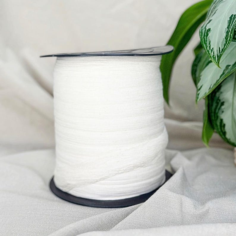 May include: A spool of white elastic ribbon, tightly wound, sits on a black base. The ribbon is textured and appears soft. A dark, thin disc sits on top of the spool. Green leaves are visible in the background.