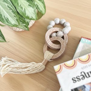 Puede incluir: Un mordedor de madera con dos círculos entrelazados, una borla y una cadena de cuentas grises. El juguete está sobre una superficie de madera clara, con una planta en maceta y un libro en el fondo.