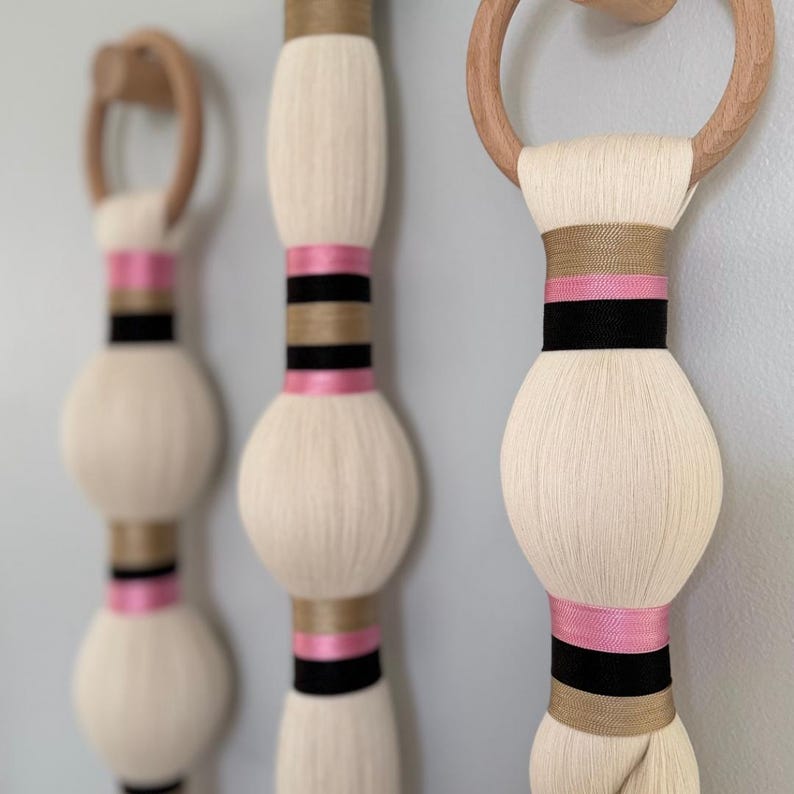 May include: Three decorative wall hangings with a cream-colored, bulbous design. Each piece features pink, black, and tan ribbon accents. The top of each hanging has a wooden ring for easy display. A modern, minimalist aesthetic.
