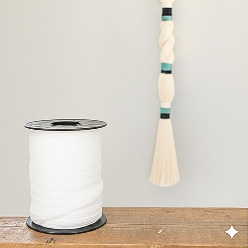 May include: A spool of white tulle fabric sits on a wooden surface. The tulle is tightly wound on a black plastic spool. A decorative hanging piece with a braided design and teal accents is also visible.
