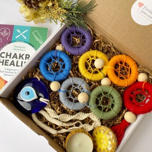 Puede incluir: Una caja de regalo llena de artículos de curación de chakras, que incluyen un libro titulado "Chakra Healing: A Beginner's Self-Healing Guide to Balance", un amuleto azul de mal de ojo, un conjunto de siete atrapasueños de colores arcoíris, un cordón de macramé blanco, una vela pequeña y un portavelas amarillo.