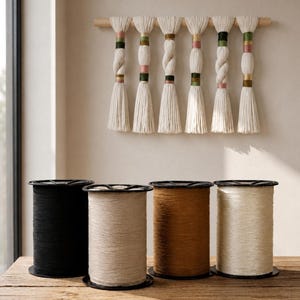 May include: A wooden wall hanging with six off-white yarn tassels, each with decorative colored bands of pink, green, and gold. Below, four spools of yarn in black, beige, and brown sit on a wooden surface, offering a range of colors for crafting projects.