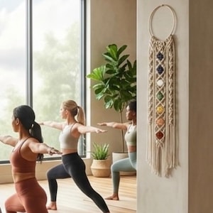 May include: A macrame wall hanging featuring a circular wooden hoop at the top and long, cream-colored cotton cords. Seven colorful, polished gemstone beads are arranged vertically in the center, representing the chakra colors in a diamond pattern.