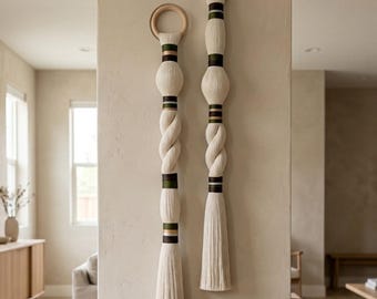 May include: Two vertical wall hangings featuring cream-colored cotton cord wrapped around wooden rings. The designs include sections of twisted cord, smooth wrapped segments, and horizontal bands in dark green and metallic gold accents.
