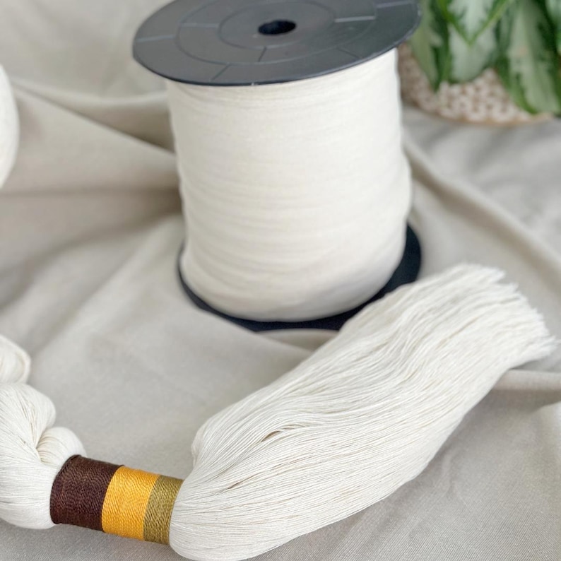 May include: A spool of white thread sits next to a bundle of the same thread, with a decorative band of brown, yellow, and green. The thread is on a neutral-colored fabric, suggesting a craft or textile theme.
