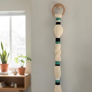 May include: A cream-colored wall hanging with a wooden ring at the top. The hanging features a series of cream-colored, bulbous shapes and a braided section, accented with black and teal bands. The piece is mounted on a light gray wall.