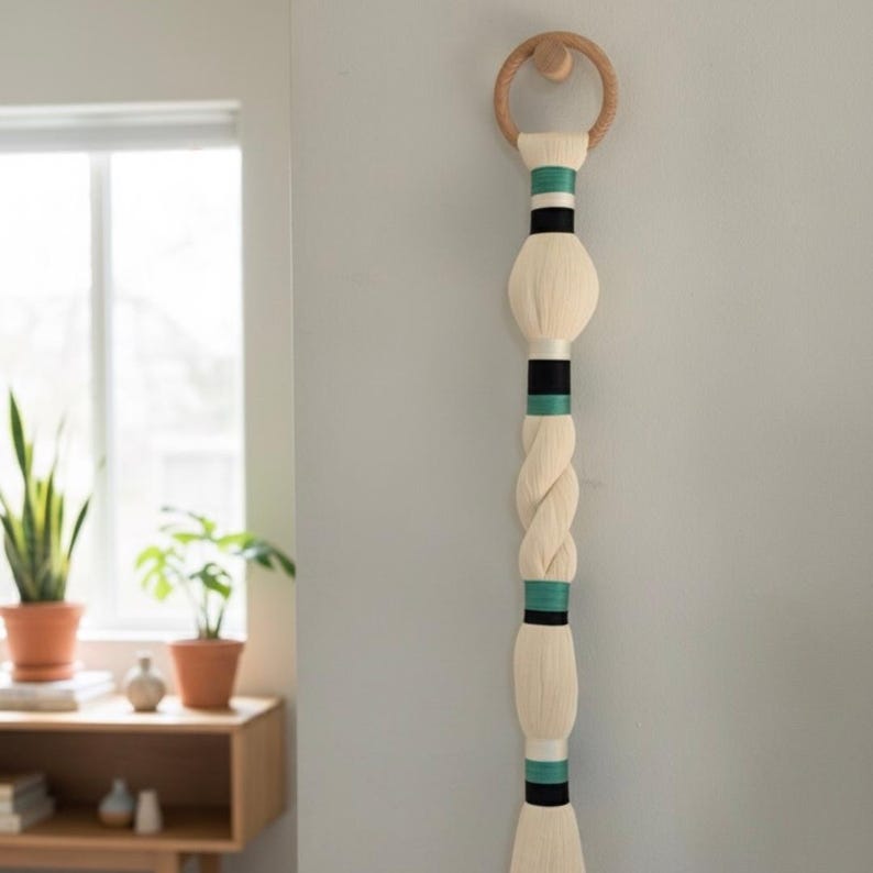 May include: A cream-colored wall hanging with a wooden ring at the top. The hanging features a series of cream-colored, bulbous shapes and a braided section, accented with black and teal bands. The piece is mounted on a light gray wall.