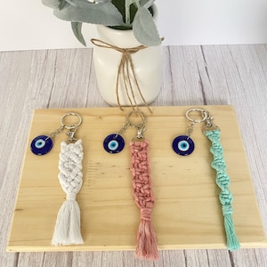 Puede incluir: Cuatro llaveros con dijes de ojo turco azul y borlas de macramé de diferentes colores. Las borlas son blancas, rosadas y verde claro.