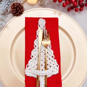 Könnte beinhalten: Ein weißer gehäkelter Weihnachtsbaum-Serviettenring mit einer goldenen Gabel und einem Messer auf einer roten Serviette. Der Serviettenring befindet sich auf einem goldenen Unterteller.