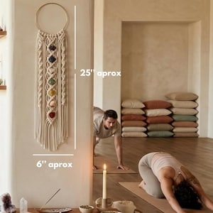 May include: A cream-colored macrame wall hanging featuring a circular gold hoop at the top and seven vertically aligned colorful gemstones in the center. The piece measures approximately 25 inches in height and 6 inches in width.