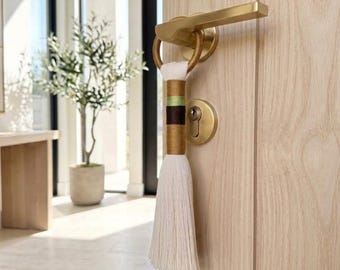 May include: A decorative tassel hanging from a brushed gold door handle on a light wood door. The tassel features a cream-colored cord with horizontal bands of metallic gold, light green, and dark brown, ending in a long cream-colored fringe.