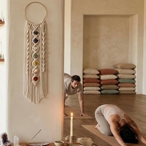 May include: A wall hanging featuring a circular gold hoop with off-white macrame cord knotted in intricate patterns. Seven polished, colorful stones are vertically aligned in the center, representing the chakra colors: purple, blue, green, yellow, orange, and red.