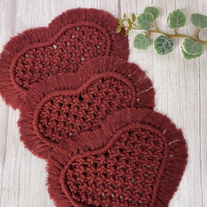 Puede incluir: Tres posavasos de crochet en forma de corazón con flecos en un color rojo burdeos oscuro. Los posavasos están apilados uno encima del otro sobre una superficie de madera blanca.