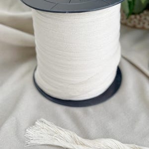 May include: A large spool of off-white macrame cord sits on a black plastic base. A length of the same cord is unraveled and lies in front of the spool. The cord is tightly wound and appears to be made of a natural fiber.