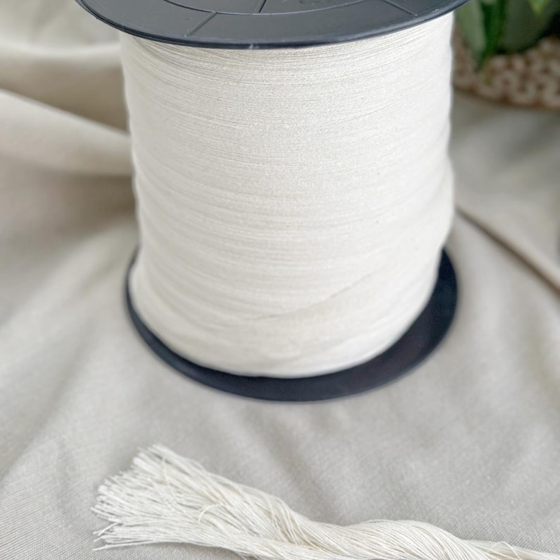 May include: A large spool of off-white macrame cord sits on a black plastic base. A length of the same cord is unraveled and lies in front of the spool. The cord is tightly wound and appears to be made of a natural fiber.