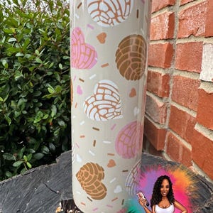 May include: A beige tumbler featuring a pattern of concha bread shapes in brown, pink, and white, along with heart shapes and sprinkles. The tumbler has a silver lid and the text "Sprinkled with Sarcasm".