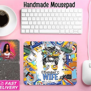 May include: A handmade mousepad with a colorful design featuring a woman with sunglasses and the text "Plumber's Wife." The mousepad is surrounded by tools and machinery graphics. The words "Handmade Mousepad" are at the top.