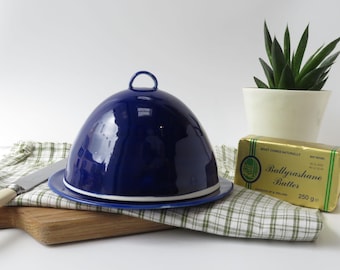 May include: A blue ceramic butter dish with a white rim and a domed lid. The dish is sitting on a checked tablecloth with a stick of butter and a knife.