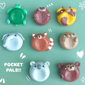 Puede incluir: Una colección de ocho pequeños platos de cerámica redondos con forma de animal etiquetados como POCKET PALS!!. Las figuras incluyen una tortuga verde, un oso marrón, una vaca amarilla, un elefante gris, un panda rojo, una foca bronceada, un koala azul claro y un mono marrón.
