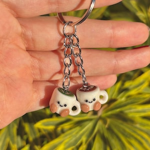 May include: A silver keyring featuring two miniature ceramic coffee cup charms. One cup has a green leaf design, the other a brown coffee design. Both have smiling faces and small tan feet.