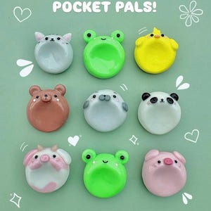 Może przedstawiać: Dziewięć kolorowych, okrągłych figurek Pocket Pals! w kształcie zwierząt, w tym kota, żaby, kurczaka, niedźwiedzia, foki, pandy, krowy, żaby i świni. Tło jest jasnozielone z białymi elementami dekoracyjnymi i tekstem "POCKET PALS!"