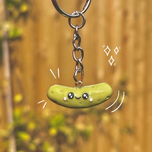Op de afbeelding: Een sleutelhanger in de vorm van een groene augurk met een lachend gezicht en zwarte ogen. De sleutelhanger is bevestigd aan een zilveren ketting en ring. De achtergrond is een wazig uitzicht op een houten hek en groen gebladerte.