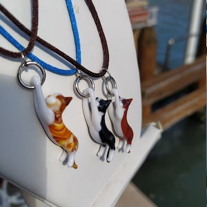 Peut inclure: Trois pendentifs en verre en forme de chat suspendus à des cordons marrons et bleus. Les chats sont dans différentes poses et sont orange et blanc, noir et blanc et marron et blanc.