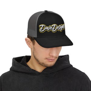 May include: Black and grey trucker cap featuring the text "Dave Designs" in yellow and white script. The cap has a curved peak and a mesh back.