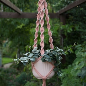 May include: A pink macrame plant hanger, suspended from a wooden ring. The woven design holds a white planter filled with green plants. The macrame has a tassel at the bottom. The background is a blurred outdoor scene with green foliage.