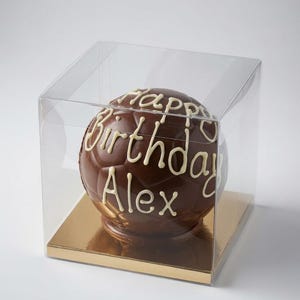 Op de afbeelding: Een bolvormige voetbal van melkchocolade met witte glazuurtekst die Happy Birthday Alex leest. De chocoladebal wordt gepresenteerd in een doorzichtige vierkante plastic displaydoos met een goudkleurige bodem voor een helder zicht.