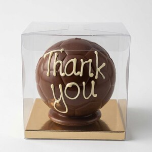 Op de afbeelding: Een bolvormige voetbal van melkchocolade met de woorden Thank you in wit glazuur op de voorkant. De chocoladebal staat op een gouden vierkante voet in een doorzichtige, vierkante plastic doos voor presentatie.