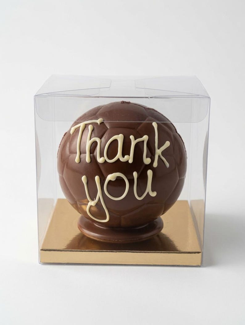 Op de afbeelding: Een bolvormige voetbal van melkchocolade met de woorden Thank you in wit glazuur op de voorkant. De chocoladebal staat op een gouden vierkante voet in een doorzichtige, vierkante plastic doos voor presentatie.