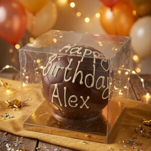 Op de afbeelding: Een bolvormige chocolade voetbal in een vierkante, doorzichtige plastic geschenkdoos. Het chocoladeoppervlak is voorzien van witte glazuurtekst Happy Birthday Alex en staat op een gouden vierkante ondergrond.