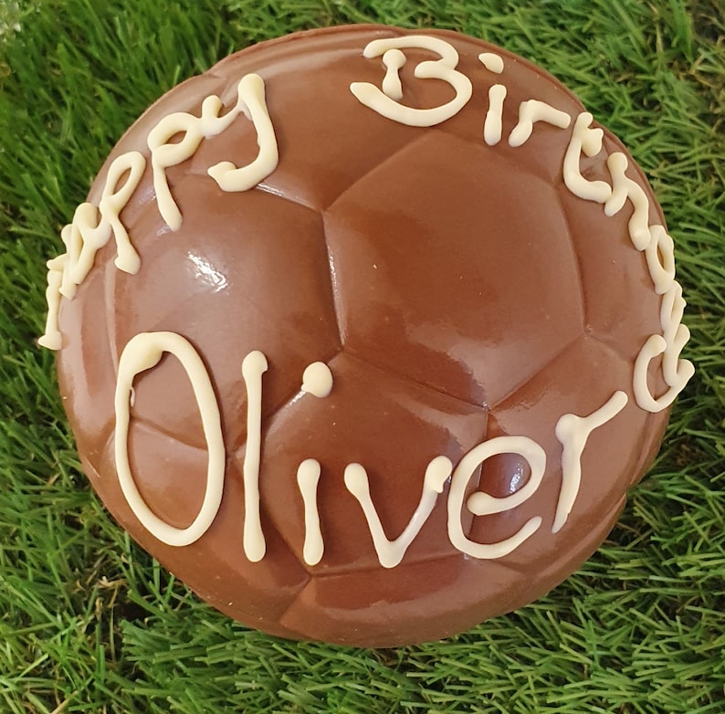 Op de afbeelding: Een bolvormige voetbal van melkchocolade met een gegoten zeshoekig patroon. Het oppervlak is versierd met witte glazuurtekst die Happy Birthday Oliver leest, geschreven in een cursief handschrift over de bovenkant van de chocoladebal.