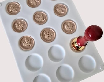 May include: White silicone mold with 12 round cavities for wax seals. The mold has a leafy design in each cavity. A red and gold wax seal stamp is placed in one of the cavities.