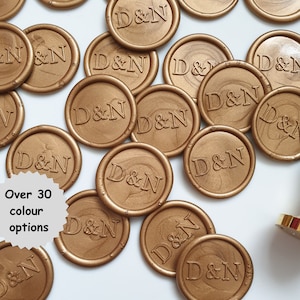 May include: A close-up of multiple gold wax seals with the initials "D&N" embossed on each. The seals are arranged in a random pattern on a white surface. There is a gold wax seal stamp in the bottom right corner of the image. The text "Over 30 colour options" is visible in the lower left corner of the image.