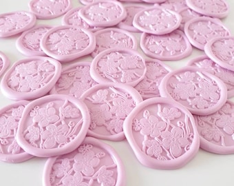 May include: A collection of light pink wax seals with a floral design. The seals are round with a raised floral pattern in the center. The seals are scattered on a white surface, with a few on a gray envelope.