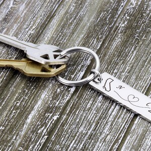 May include: Silver keychain with a silver key ring and two keys. The keychain has a silver charm with the outlines of California and North Carolina, a dotted line connecting the two states, and a heart in the middle of the line. The charm is engraved with the letters "N" and "C".