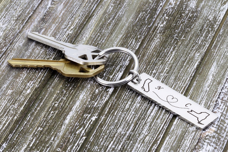 May include: Silver keychain with a silver key ring and two keys. The keychain has a silver charm with the outlines of California and North Carolina, a dotted line connecting the two states, and a heart in the middle of the line. The charm is engraved with the letters "N" and "C".