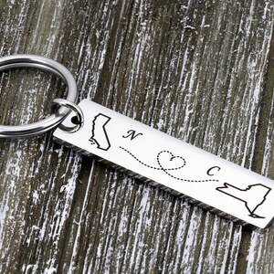May include: Silver keychain with a map of California and New York connected by a dotted line with a heart in the middle. The keychain says "N" next to California and "C" next to New York.