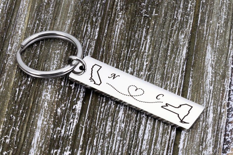 May include: Silver keychain with a map of California and New York connected by a dotted line with a heart in the middle. The keychain says "N" next to California and "C" next to New York.